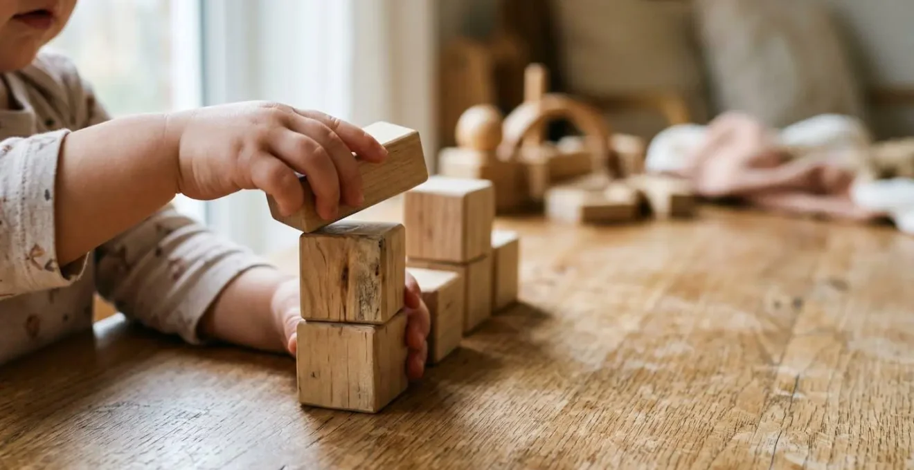 Close-up of a toddler's hands stacking simple wooden blocks on a natural wood surface, showing concentration and fine motor development in warm natural light